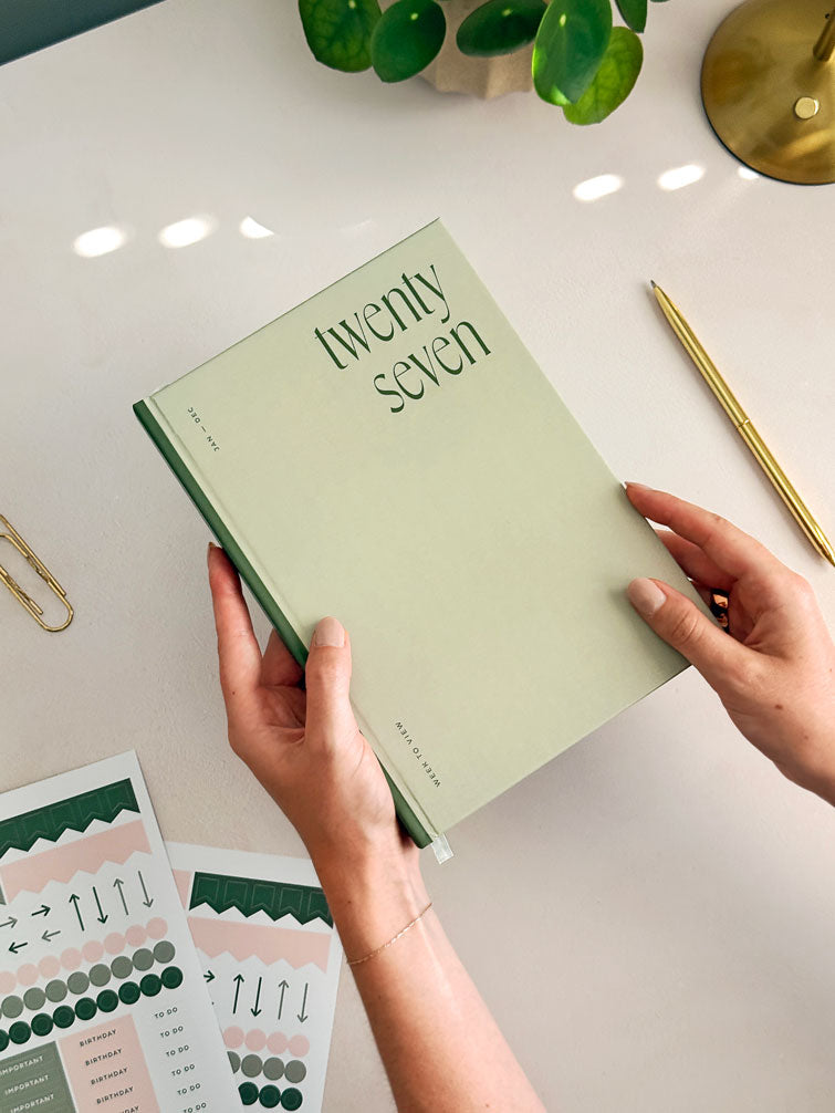 A woman holding an A5 week to view diary with soft sage green hardback cover and contrasting deep froest green spine. There&