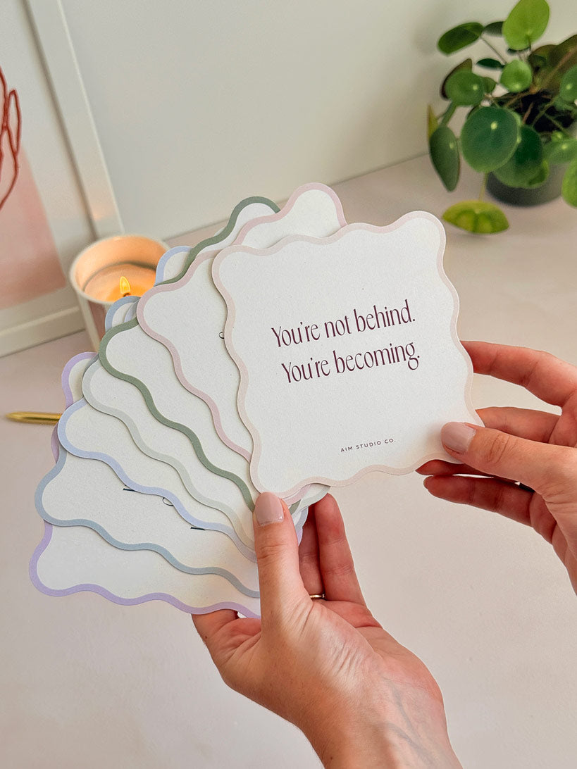 woman holding a fanned out set of positive affirmation cards that are cut into a wavy shape. The set of quote cards have a light pastel-coloured border with modern serif typography printed on reycled card with small organic flecks in the paper.