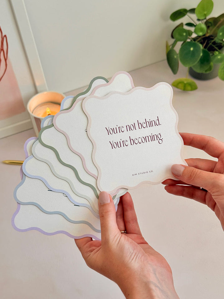 woman holding a fanned out set of positive affirmation cards that are cut into a wavy shape. The set of quote cards have a light pastel-coloured border with modern serif typography printed on reycled card with small organic flecks in the paper.