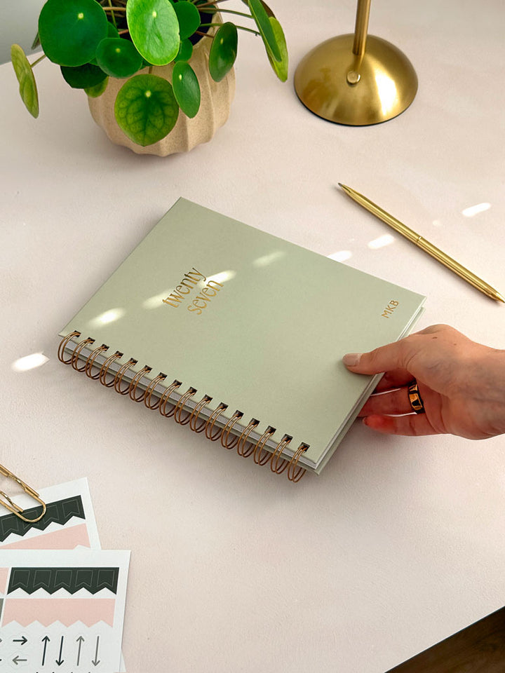 woman placing an A5 personalised diary on a desk surrounded by planner stickers and stationery. The product has spiral binding with a sage green cover and gold foiled details.