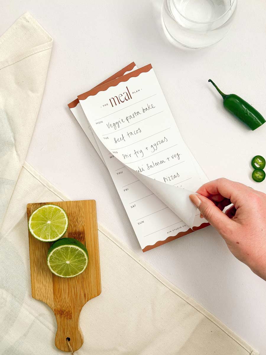Hand tearinf ogg page of weekly meal planner notepad with a cutting board and ingredients on a kitchen table
