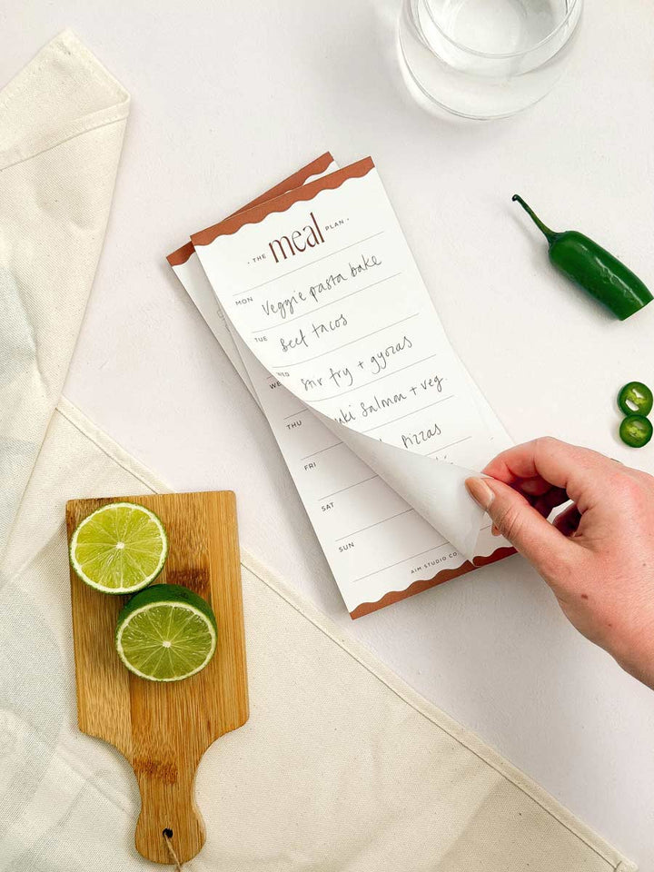 Hand tearinf ogg page of weekly meal planner notepad with a cutting board and ingredients on a kitchen table