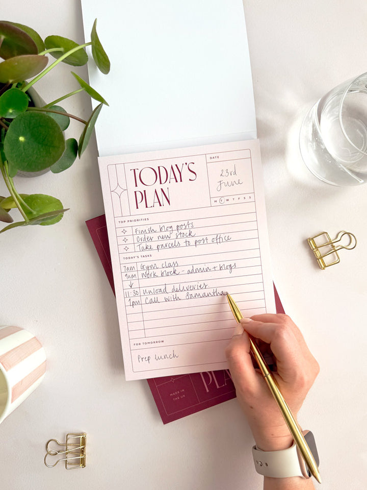 a person is writing daily to do list on the inner pages of a planner notepad with tear off sheets. The pages are pale pink and feature a structured, grid-lined design for productivity. The desk surface has gold stationery accessories, a desk plant and a glass of water.