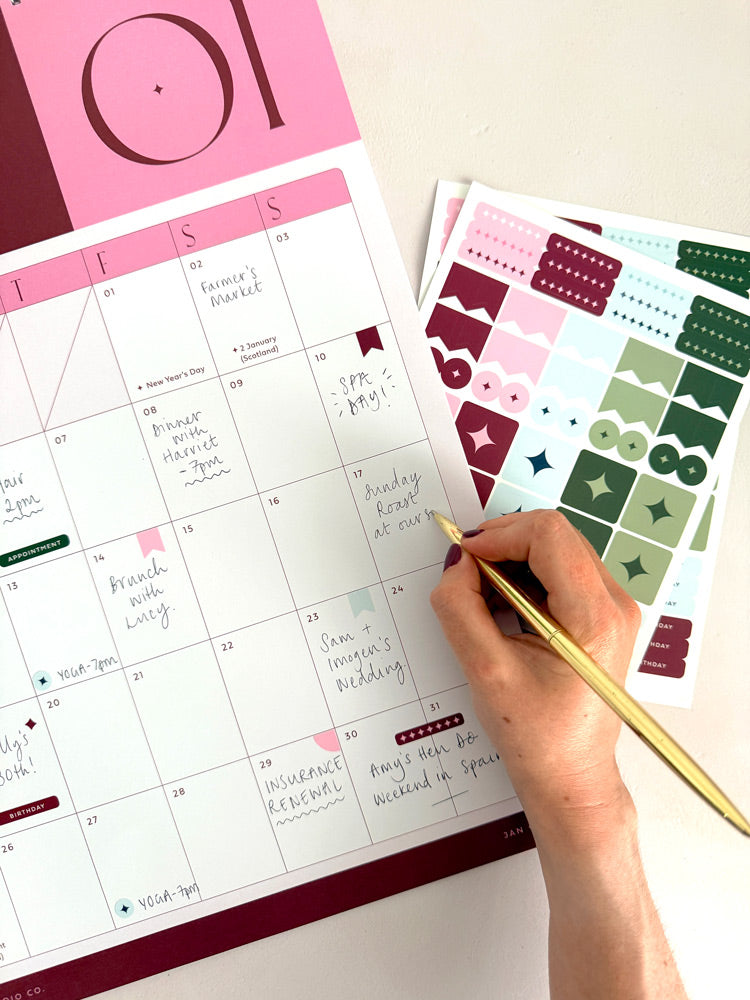 A close-up of a hand writing on a large a3 monthly planner page with a gold pen, next to sheets of decorative planner stickers in pink, green, and burgundy on a light beige surface.