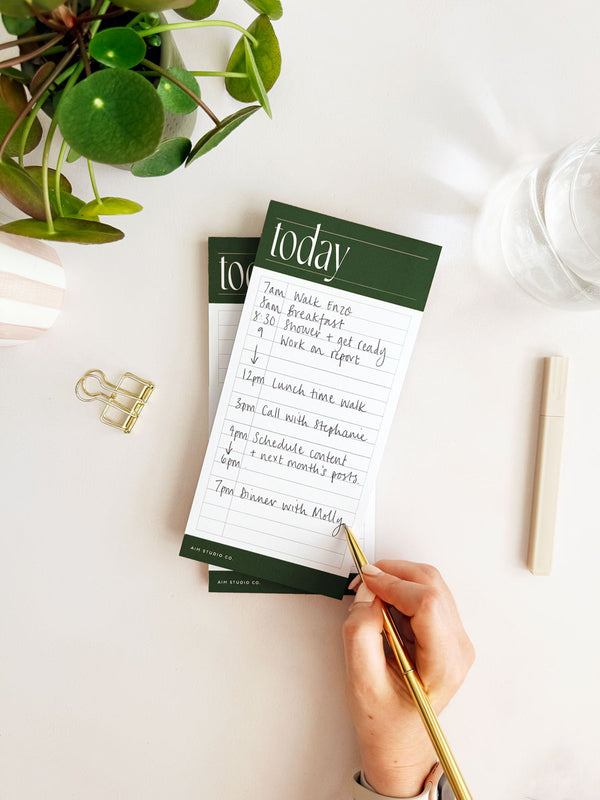 Person writing on a green and white notepad with a plant and desk items in the background
