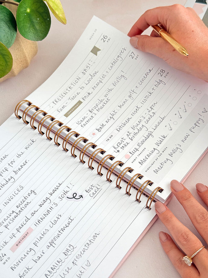 Woman turning the page of 2027 week to view diary by aim studio co. The pages are decorated with aesthetic planner stickers and have hand written plans for the week, as well as a habit tracker and box for a weekly highlight.