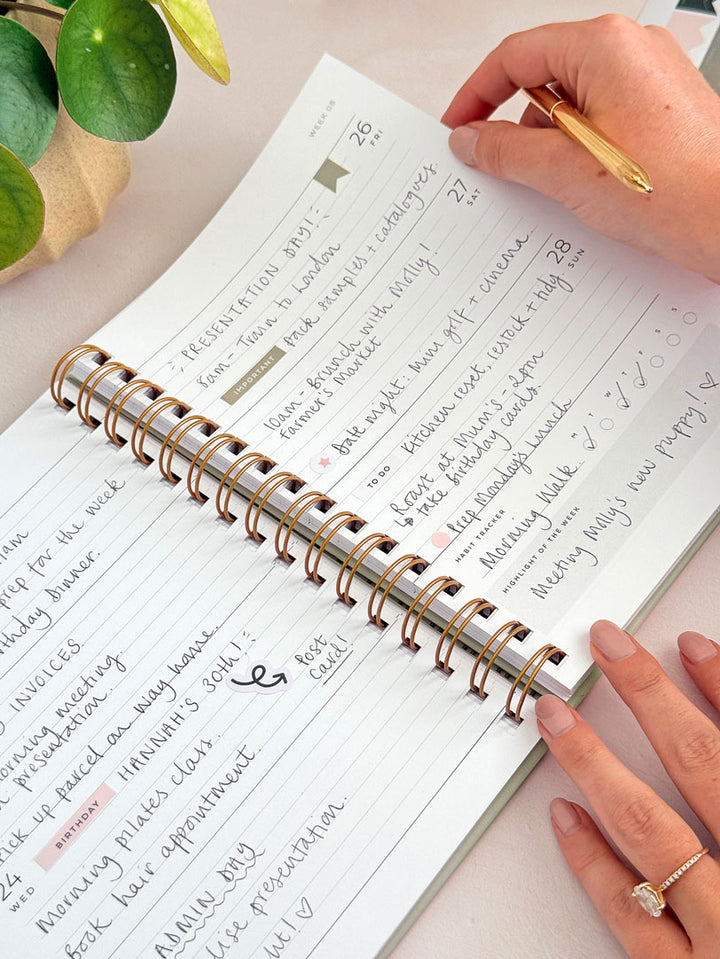Gold spiral-bound 2027 weeky planner with handwritten plans across the pages. The diary layout shows lined sections with UK bank holidays and is decorated with sage green planner stickers.