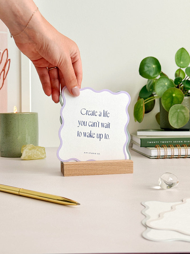 wavy-shaped card with positive quote and lilac border displayed in a wooden holder on a desktop. A woman is changing the card to reveal a new daily affirmation. Styled in a modern office desk space with candle and crystals.