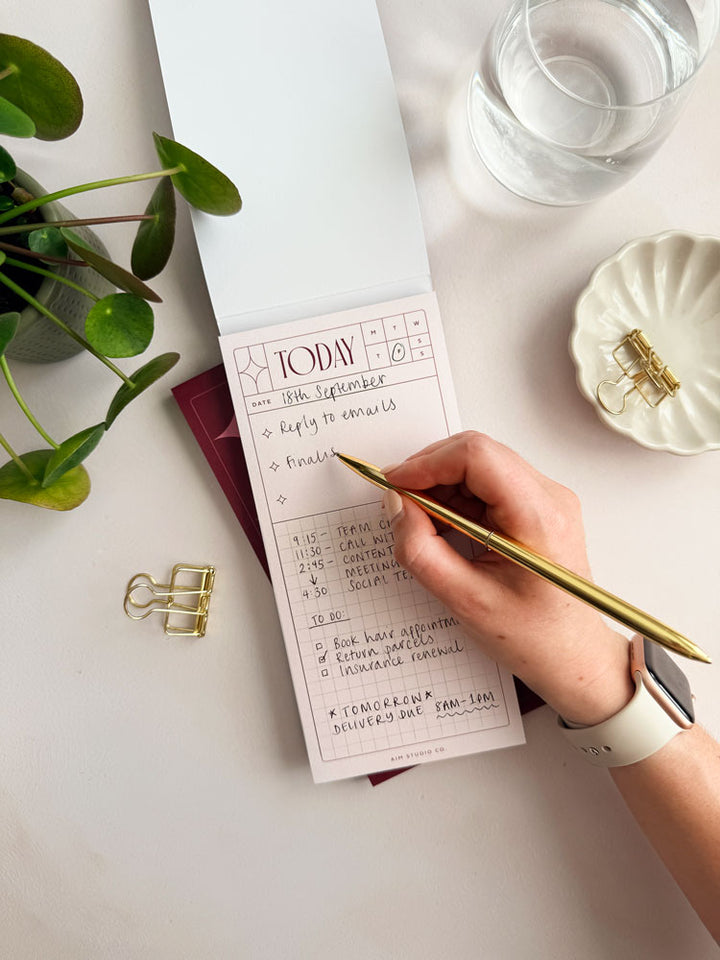 woman writing daily task list inside slim notepad with pale pink pages and tear-off sheets. The planner has space for top priorities each day and a grid-lined planner below for to do list and time blocking.