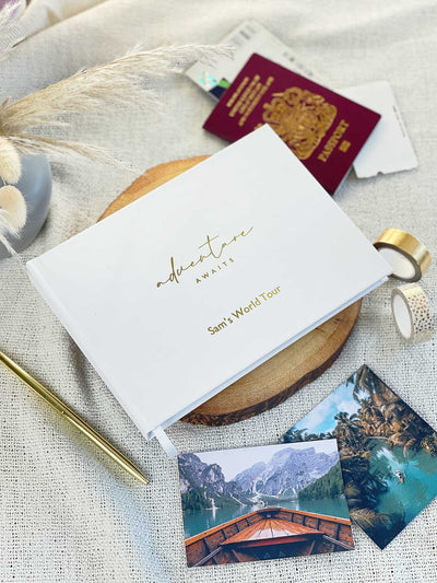 white memory album with a UK passport and travel photographs surrounding a white and gold hardcover memory book with adventure awaits on the cover and personalised gift message to a couple on a world tour
