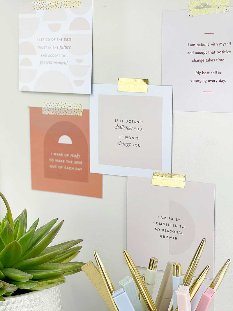 Positive affirmation cards taped on a wall with gold washi tape above aesthetic office desk with plants and gold accessories in a pen pot