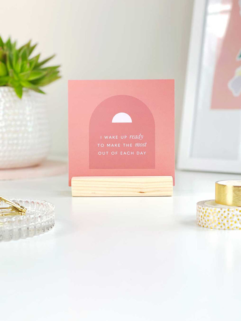 A coral coloured positive quote card stood in a wooden holder to display a set of positive affirmation cards